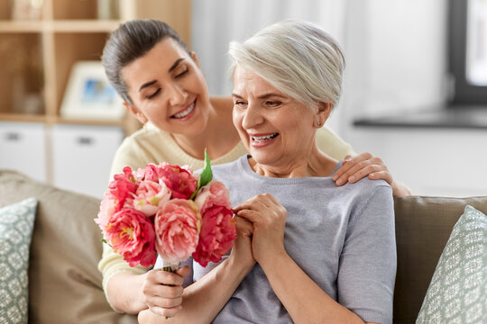 family, mother's day and birthday concept - happy smiling adult daughter giving flowers to her senior mother at home