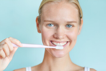 Happy young woman brushing her teeth white while looking at the camera smiling with beautiful eyes. Isolated against a blue background.