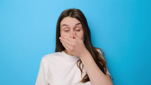 Shocked brunette girl isolated on blue studio background looking at camera with big eyes shocked expression, closing mouth zipping lips to keep terrible secret. People, emotions and unexpectedness