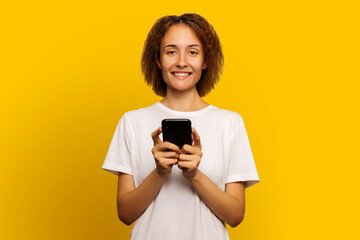 Fototapeta premium Portrait of a woman with curly hair in a white tank top holding a smartphone and smiling while looking at the camera on an isolated yellow background. Woman writes sms. Good news