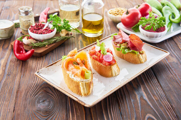 Bruschetta with various toppings, variety of small sandwiches with salmon red fish, fresh vegetables, tomatos and herbs on wooden table, diet food ingredients background.