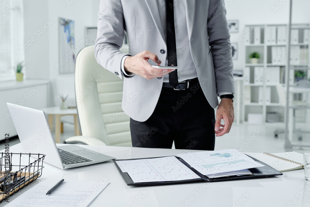 Poster unrecognizable businessman standing at desk with laptop and taking photos of documents while prepari