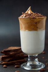 Glass of trendy fluffy creamy dalgona or whipped instant coffee with milk, coffee beans and chocolate on the dark background. New popular food and drink trend concept. Copy space.