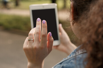 Young curly girl using white tablet pc outside. Woman sitting in the city park and holding modern gadget. Focus on hand with ring. Mock up picture of lady with portable e-book reader.