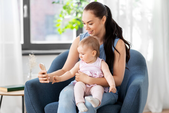 family, child and motherhood concept - portrait of happy smiling mother and little baby daughter with smartphone sitting in chair at home