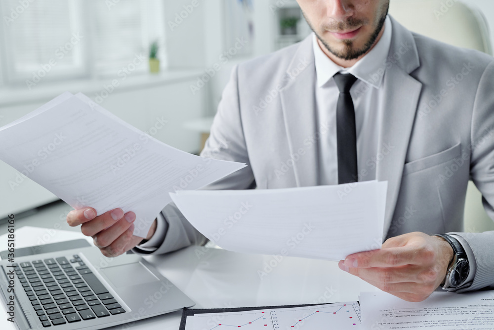 Poster close-up of concentrated young bearded manager sitting at desk in office and analyzing document cont