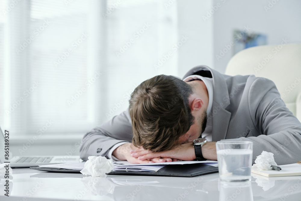 Poster exhausted young businessman lying on table with papers and crumpled napkins while suffering from hea