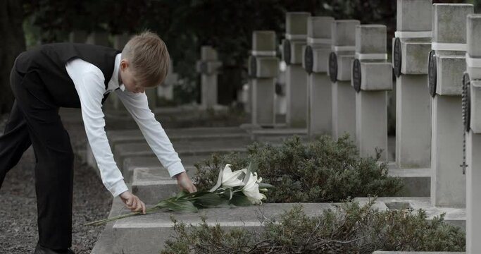 Side view of young boy putting white lily flowers on gravestone of his father. Teenager kid honoring his dad soldier at cemetery. Concept of memorial day