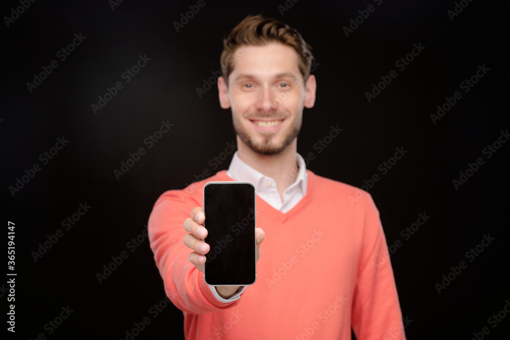 Poster portrait of sales manager in orange sweater presenting new smartphone and showing it at camera, blac
