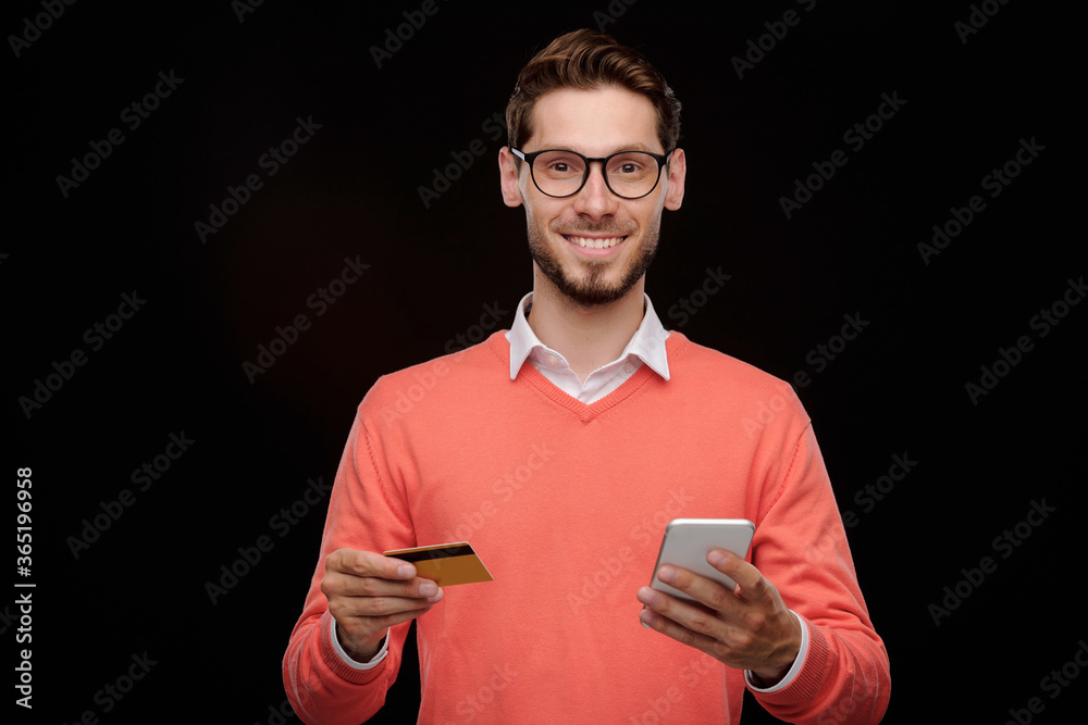 Poster Portrait of cheerful handsome young man with stubble standing with credit card and smartphone and using online transaction service