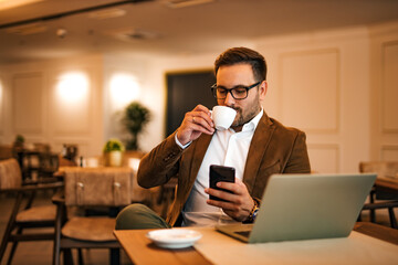 Successful businessman drinking cup of coffee at the cafe. Sitting at the table with laptop and using smart phone.