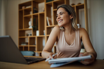 Fototapeta premium Portrait of a charming smiling university student, sitting at table with laptop and note pad and looking away.