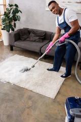African american cleaner in uniform and rubber gloves smiling at camera while cleaning carpet with...