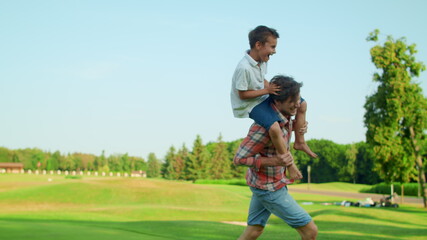 Positive man and boy running in field. Happy father carrying son on shoulders