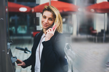 Smiling attractive female traveler using call box telephone during city tour standing on urban settings background, positive hipster girl satisfied with good connection talking on phone being in trip