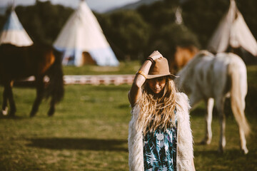 A blonde bohemian woman with a hat outdoors in a meadow with horses background