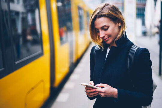 Positive blonde woman holding cellular updating information about city transport on web page, smiling hipster girl satisfied with online ticket service paying for electric transport via smartphone