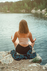 Young woman practicing yoga in the nature, sitting on the stone back to the camera and meditation, lake, active life concept, summer time