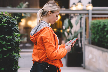 Side view of young female using mobile phone data internet connection while standing on city street. Caucasian hipster girl in orange jacket reading text message via chat application on smartphone