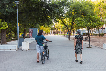 Man dragging bicycle and woman standing with mask, talking quietly and walking in an urban park while maintaining social distance to prevent the spread of the coronavirus in Spain. Selective focus.