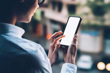 Rear view from shoulder of female person touching with finger copy space screen of modern smartphone standing outdoors near bokeh lights background. Woman using mobile phone applications with internet