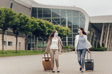 Two happy women in protective masks after coronavirus quarantine with suitcases go to the airport.