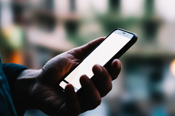 Side close up view of male person holding modern smartphone with mock up copy space screen while standing against bokeh background. Man using mobile phone device applications with internet connection