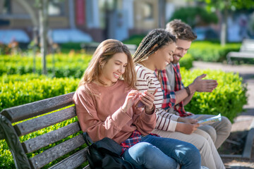 Girl with smartphone and girlfriend with guy with map on bench