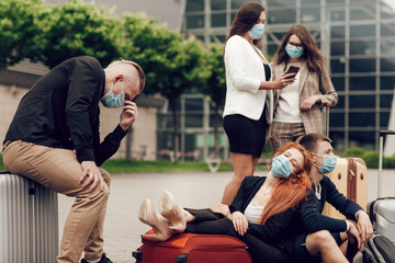 Obraz premium Close up portrait of a five friends waiting for the flight, sitting on the asphalt, using mobile phones. Three girls and two boys in protective masks with suitcases go on a trip