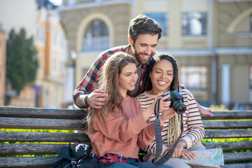 Guy is standing two girls sitting with camera in hands.