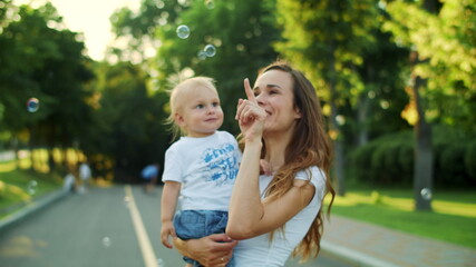 Mother holding toddler on hands. Woman and boy looking at soap bubbles on street