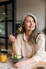 Woman sitting at kitchen indoors at home while eating salad.