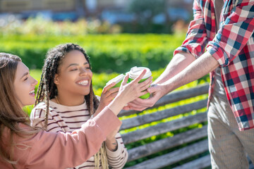 Obraz premium Two smiling girls taking glasses of coffee from male hands