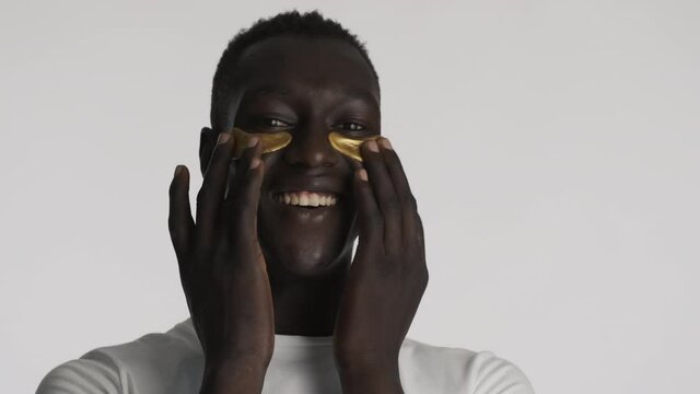 Attractive cheerful African American guy with gold eye patches joyfully posing on camera over gray background. Facial treatment 