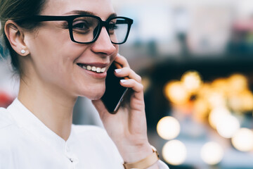 Cropped view of cheerful young woman in eyeglasses with black frame talking on smartphone device using roaming connection.Positive businesswoman calling on cellular and laughing on bokeh background