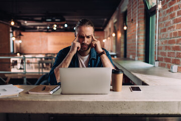 pensive businessman looking at laptop near smartphone and paper cup in office