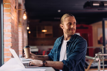 Fototapeta premium happy businessman looking away near laptop and holding disposable cup in office