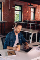 happy businessman holding paper cup and using laptop near smartphone on desk