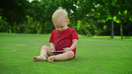 Adorable toddler holding cellphone in hands. Cheerful boy sitting on green grass