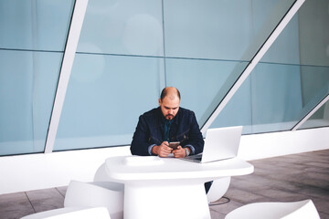 Male developer chatting on mobile phone after work on laptop computer while sitting in modern...