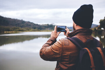 Back view of stylish hipster guy taking photo with smart mobile digital camera of a beautiful landscape during trip, male traveler shoots video on cell telephone of mountains and lake during journey