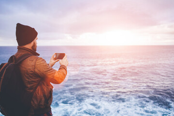 Back view of a young man tourist taking photo with cell telephone digital camera while standing in front sea with waves, hipster guy with trendy look shoots video with ocean landscape on mobile phone