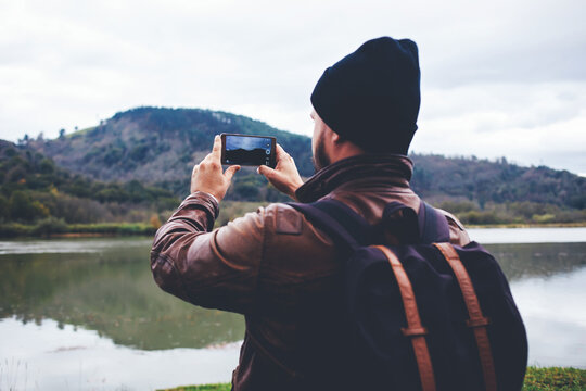 Back view of a man traveler with rucksack on his back taking photo on mobile phone camera while standing in front lake and mountains, hipster guy shoots video  of a beautiful nature on cell telephone