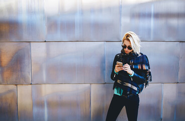 Stylish hipster girl chatting on cell telephone while standing against urban metallic background with copy space area for your text message or advertising content,young female using mobile smart phone