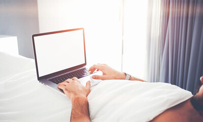 Closely of man successful freelancer is using laptop computer for remote job, while is relaxing in bed in home interior. Male is searching information on web site via net-book with copy space screen