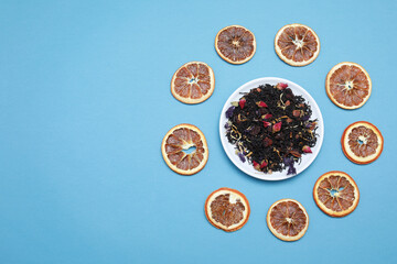 Fragrant herbal dry leaf tea with fruits and petals around white saucer with dry slice apple isolated on blue background