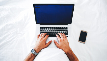 Top view of hipster guy is typing text message in social network via laptop computer, while is lying on a bed with mobile phone. Open portable net-book and cell telephone with blank copy space screens