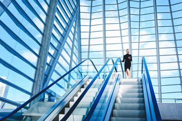 Female skilled CEO is dialing a number on mobile phone while is standing near escalator in interior with contemporary design. Woman is using cell telephone, while is moving on staircase in office