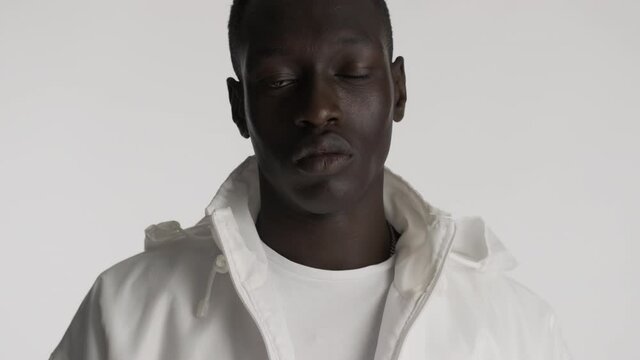 Handsome stylish African American guy in white jacket confidently taking off hood over gray background. Serious expression
