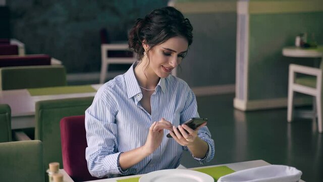 Business woman wearing formal shirt, talking online with her colleagues using smartphone in modern restaurant at luch time. Social distancing.